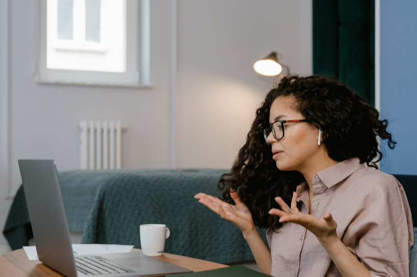 woman of colour sits at her desk engaging in a virtual meeting. She has both palms facing up as she attempts to explain something to the participants