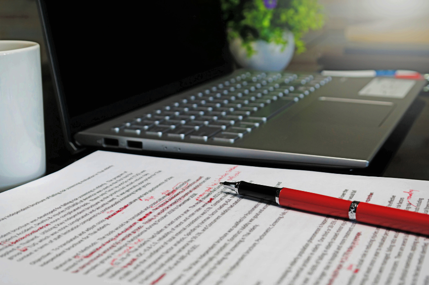 Zoomed in image of a laptop with a document next to it that has red edit notes one it.